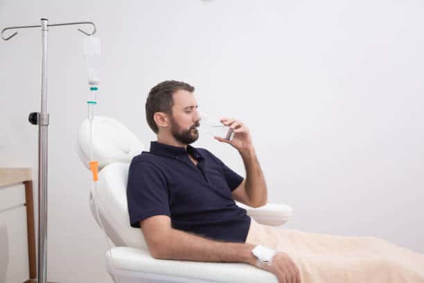 Sick, dehydrated   patient man receiving vitamin IV infusion drip and drinking glass of water in the clinic