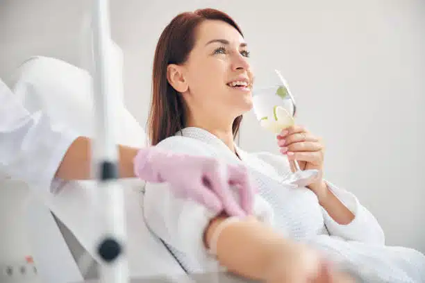 Female patient drinking a healthy beverage while sitting in medicine armchair and receiving IV infusion in wellness center.